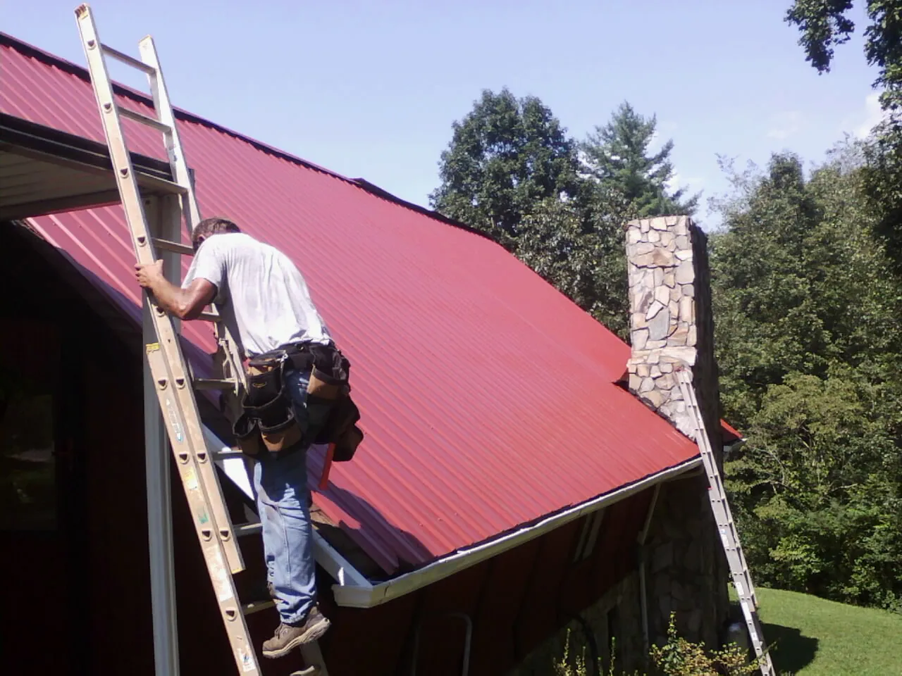 Roofer on a ladder beside a red metal roof and stone chimney — Soffit & Fascia Repair by Roofing Brothers in Earling